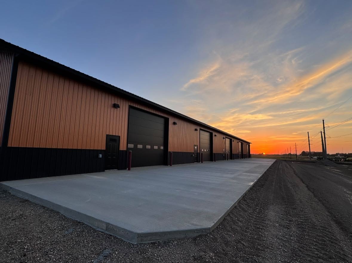 Row of heated shop condos with black overhead doors and a poured concrete approach at sunset in Rugby, North Dakota